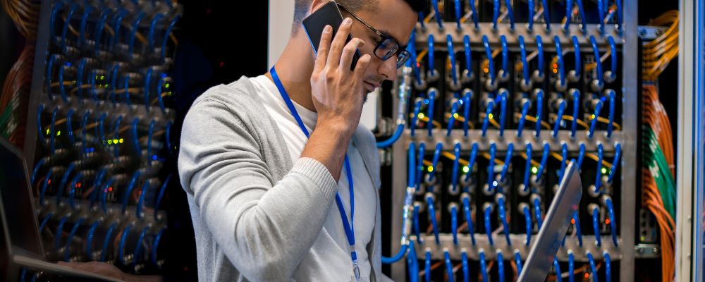 Man in a server room receiving maintenance support via a phone call and on his laptop.